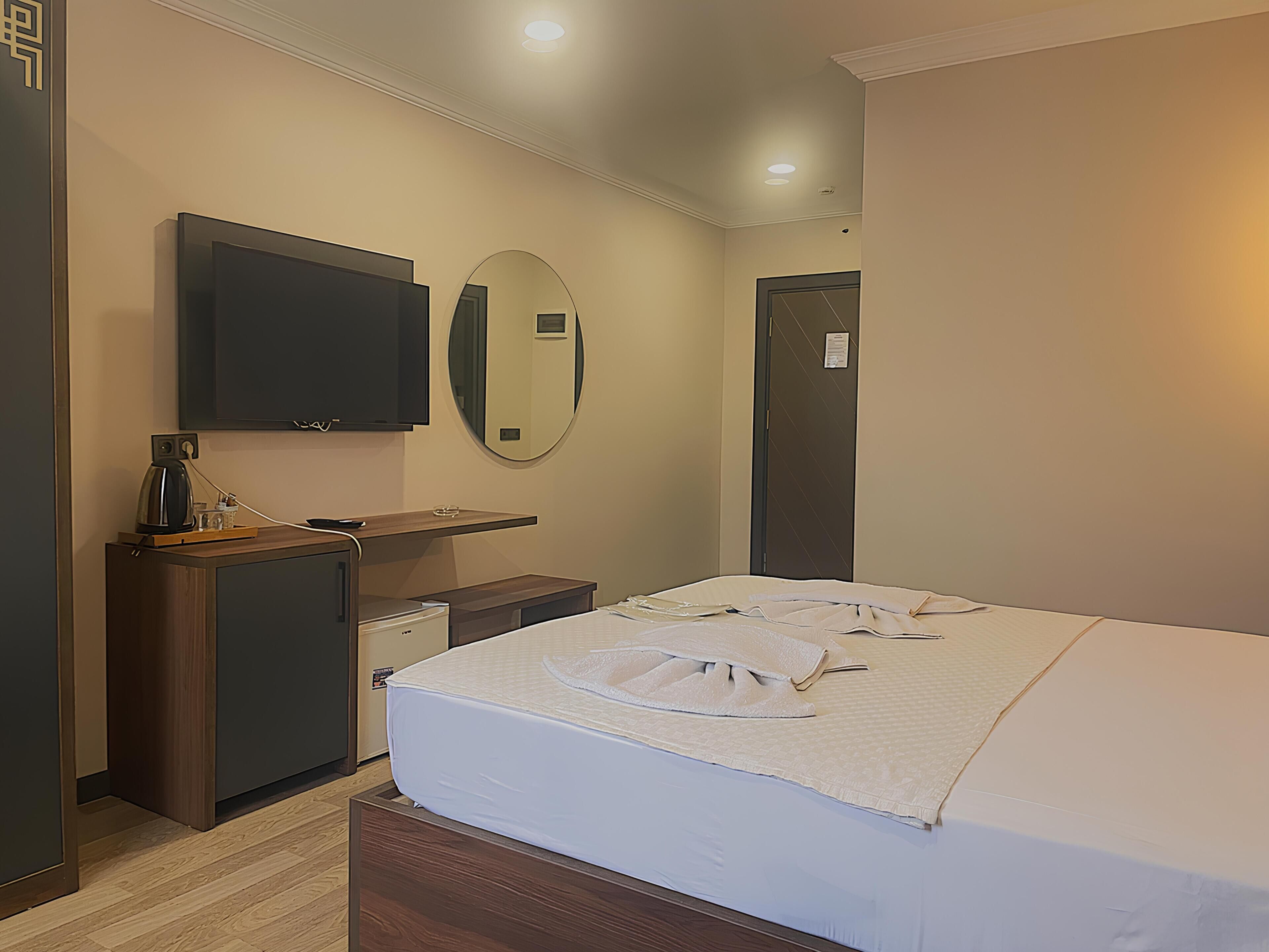 Quarto de hotel com uma cama, televisão na parede, secretária pequena com cafeteira e espelho.
