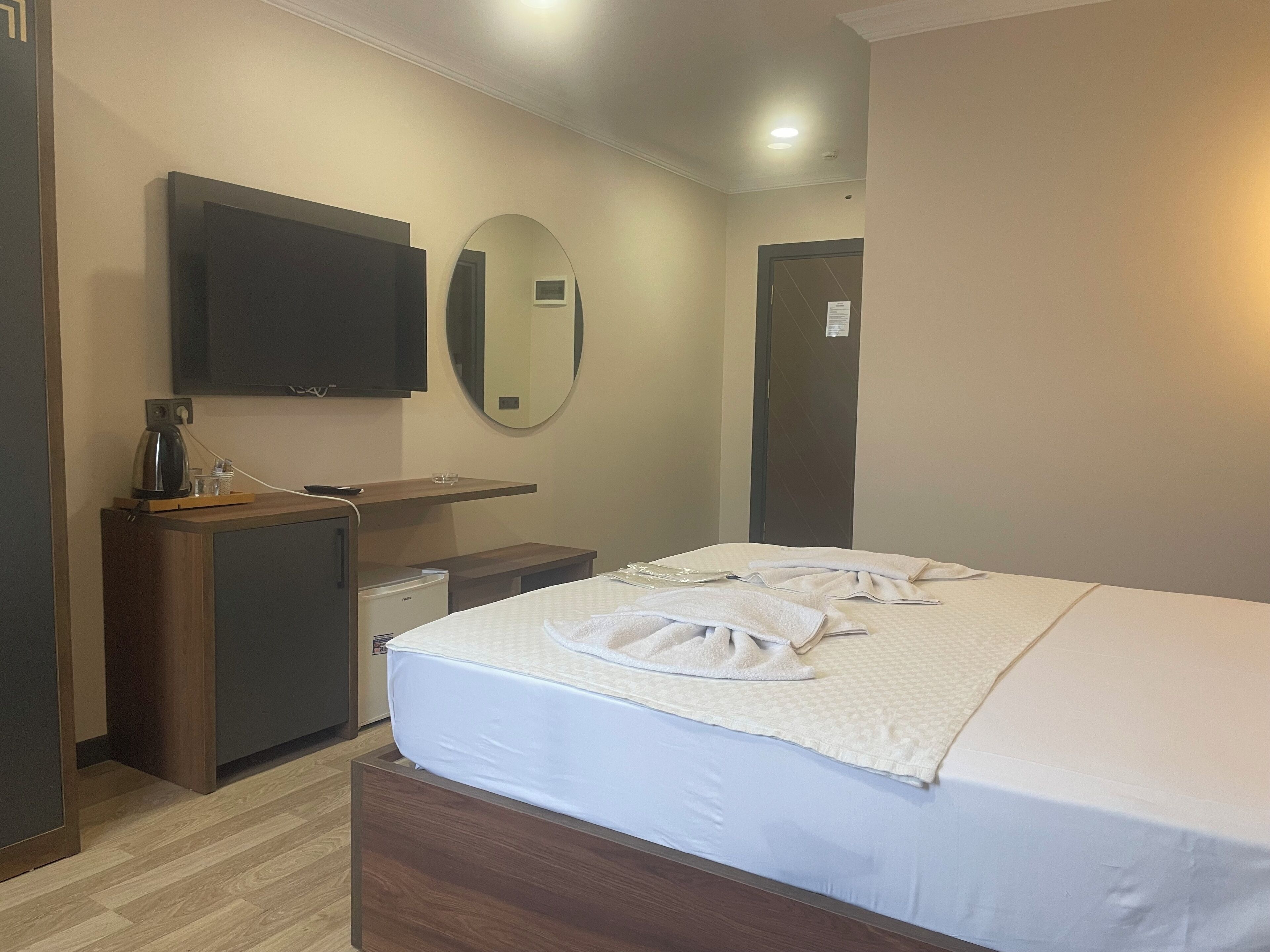 Une chambre d’hôtel avec un lit, une télévision, un petit bureau avec une bouilloire, un miroir et une porte.