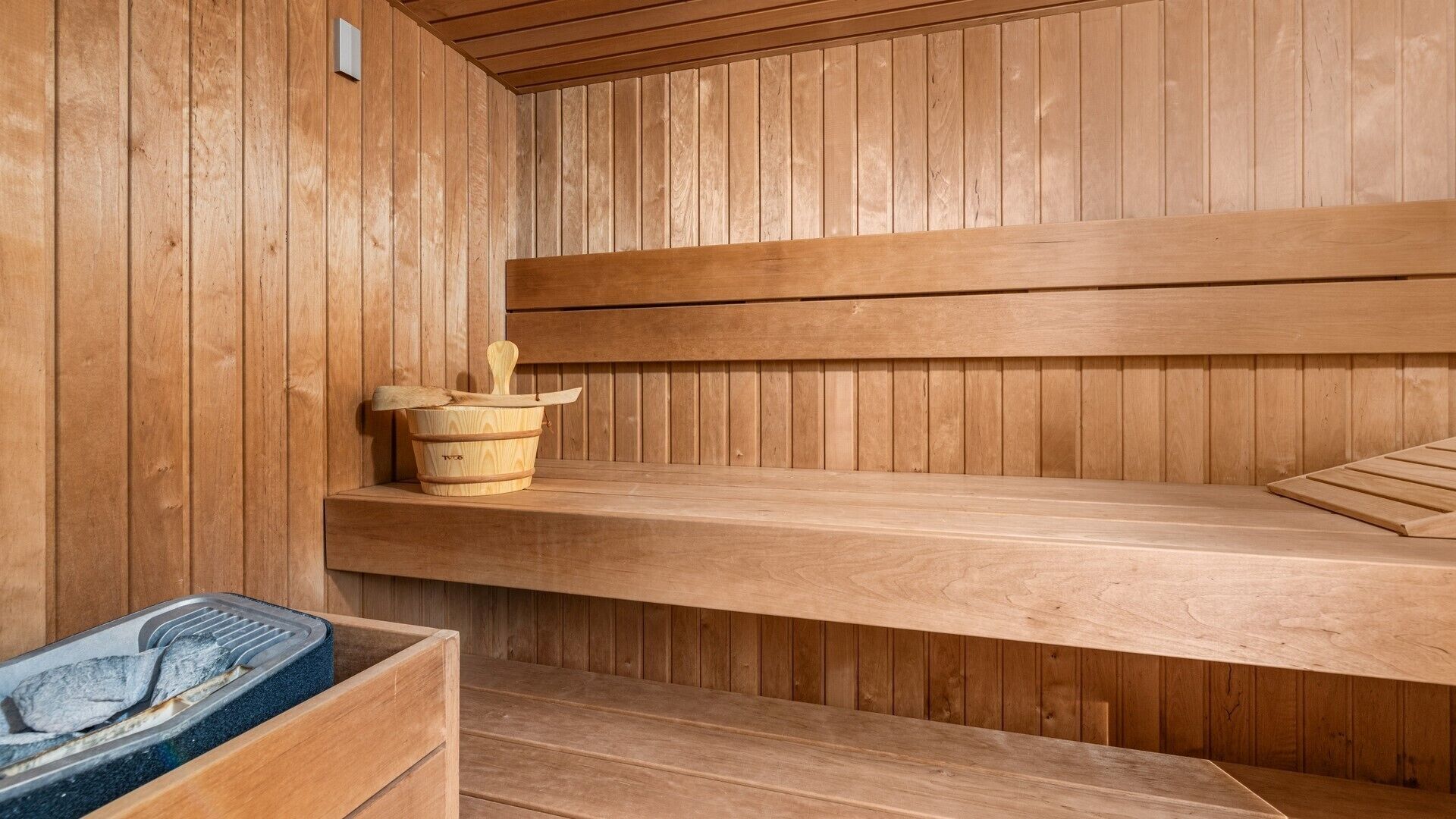 Una sauna de madera con bancos, un balde de madera y piedras de sauna visibles.
