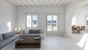 A spacious living room with a large window, a sofa, a coffee table, and a dining area with chairs. - Villa Conza Mykonos (Mykonos)