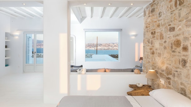 A modern bedroom with a stone wall, a large window with a city view, a built-in bench with cushions, and a wooden side table. - Villa Conza Mykonos (Mykonos)