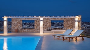 A poolside area with stone pillars, lounge chairs, and a view of a snowy landscape. - Villa Conza Mykonos (Mykonos)