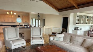 A spacious living room with a large beige sofa, two armchairs, and a leather ottoman. A kitchen with wooden cabinets and a dining area with a table and chairs is visible in the background.