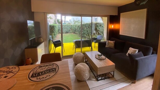 Ein modernes Wohnzimmer mit einer Couch, einem Couchtisch und Sesseln. Ein Schiebetürfenster führt zu einem Außenbereich mit einem gelben Teppich und viel Grün.