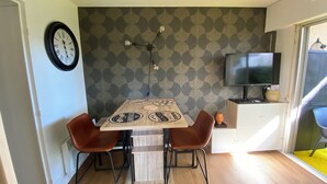 Ein Essbereich mit einem Holztisch, zwei braunen Stühlen, einer Uhr, einer Wand mit Muster und einem Fernseher sowie einem kleinen Schrank.
