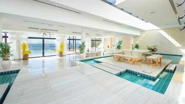 Un vaste espace intérieur abritant une piscine, avec vue sur l’océan, un coin repas avec tables et chaises, ainsi que plusieurs plantes en pot.