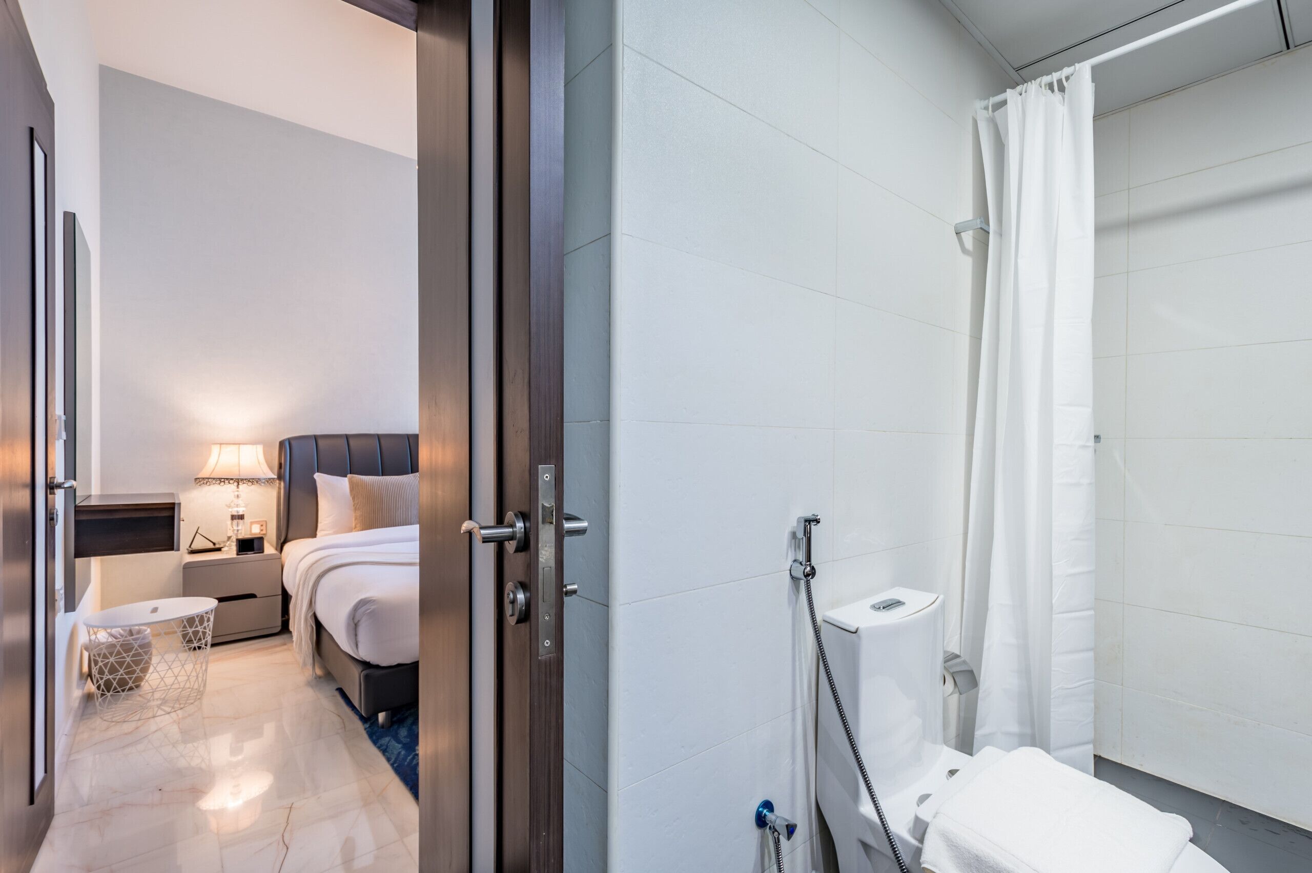Moderni hotellihuone, jossa on sänky, yöpöytä, lamppu ja suihkutila, jossa on verho.