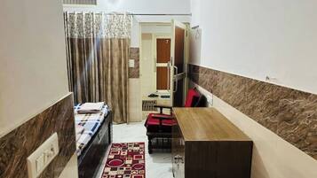 Une chambre d’hôtel étroite, avec un lit, une petite table, un banc rouge et un meuble en bois.