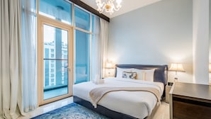 A modern hotel room with a large bed, a nightstand, a lamp, and a view of the city through floor-to-ceiling windows.