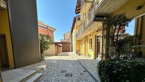 Une ruelle pavée, située entre deux bâtiments, avec un portail en métal sur la gauche et un petit arbre sur la droite.