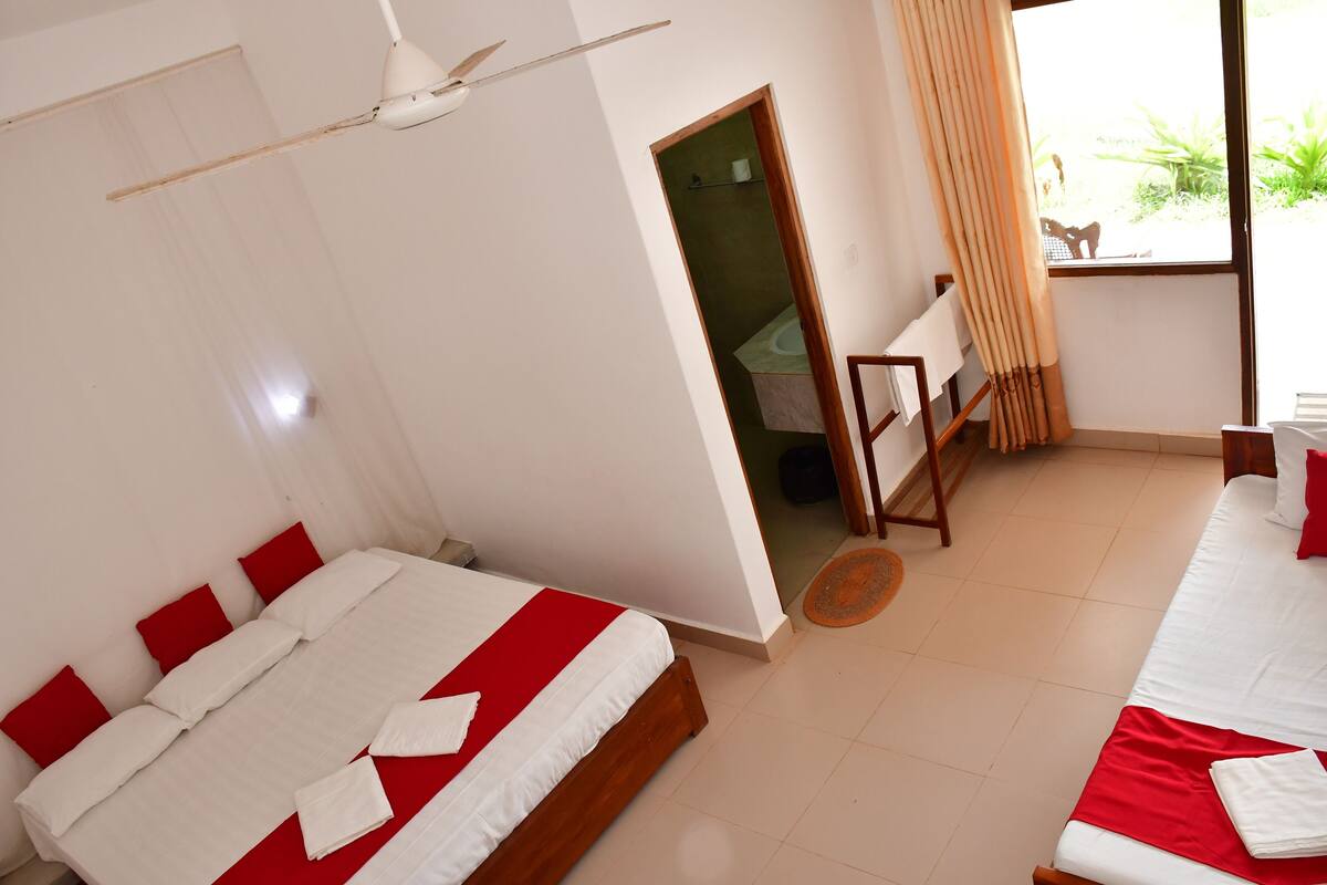 A hotel room with two beds, a ceiling fan, a window with curtains, and a view of greenery outside.