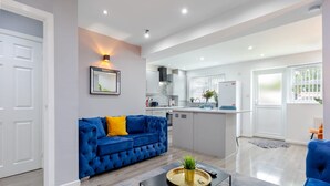 Un salon moderne avec un canapé bleu, une table basse sur laquelle se trouve un plateau, et une cuisine en arrière-plan.