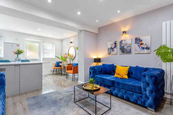 Una sala de estar moderna con un sofá de terciopelo azul, una mesa de centro y una zona de comedor con sillas naranjas.