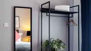 Une pièce comprenant un lit, un miroir, une étagère en métal noir avec une serviette et un cintre, et une plante en pot.