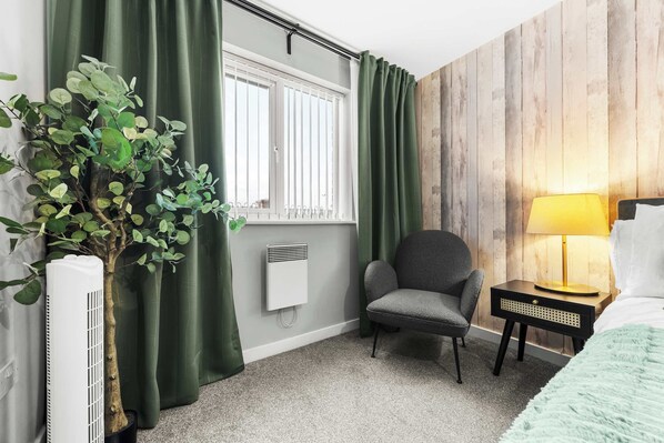Una habitación con radiador, una silla, una mesita de noche con lámpara y una ventana con cortinas.