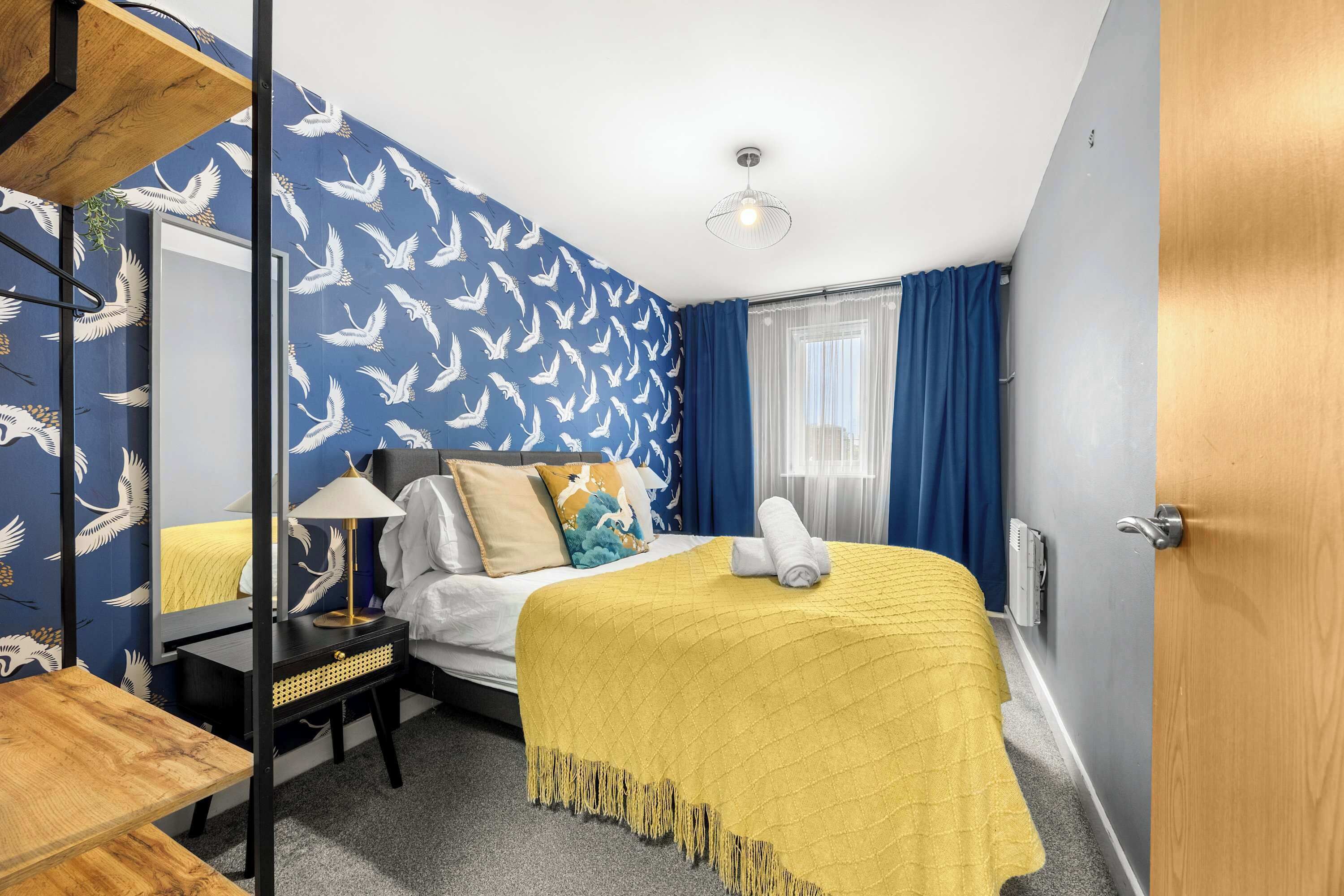 A hotel room with a bed, a bedside table with a lamp, a mirror, a window with curtains, and a door.