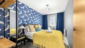 A hotel room with a bed, a bedside table with a lamp, a mirror, a window with curtains, and a door.