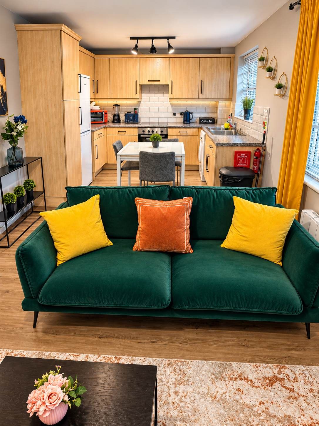 A modern kitchen with wooden cabinets, a dining table, and a sofa with yellow and orange cushions.
