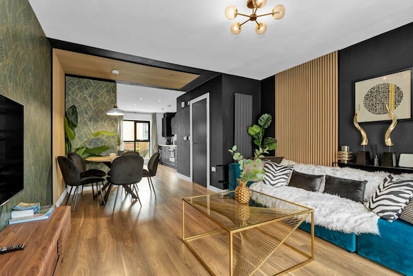 A modern living room with a wooden floor, a glass coffee table, a sofa with patterned cushions, and a dining area with chairs and a table.