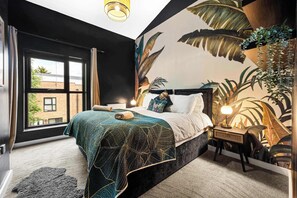 A bedroom with a large window, a bed with a green patterned bedspread, a nightstand with a lamp, and a tropical-themed wall mural. - Salford Quays Luxury Safari Retreat – Free Parking (Greater Manchester)