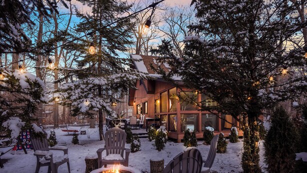 Een gezellig buitenverblijf met Adirondack-stoelen en een vuurkuil, omgeven door met sneeuw bedekte bomen en een houten hutje op de achtergrond.