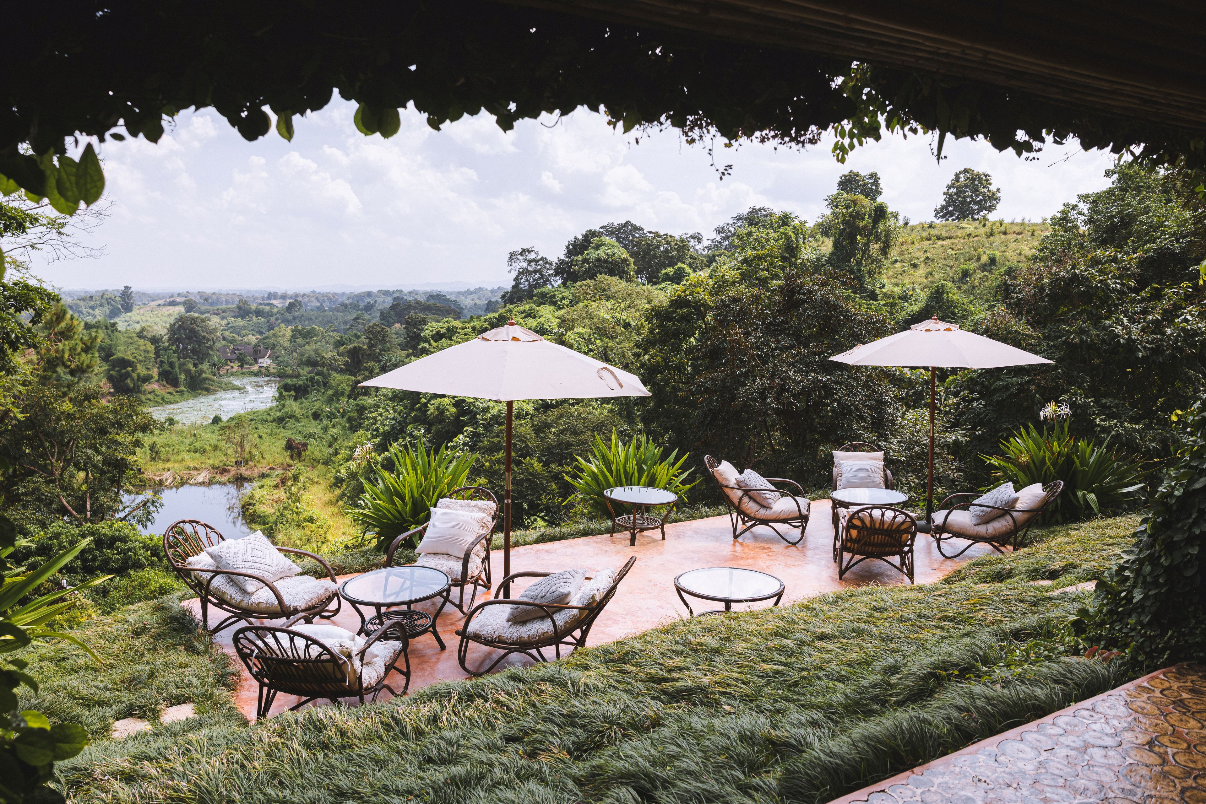 Un patio exterior con mobiliario de mimbre y sombrillas, con vistas a un paisaje exuberante con un río y colinas.