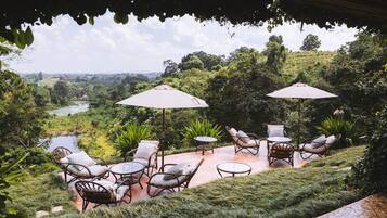 Un patio exterior con mobiliario de mimbre y sombrillas, con vistas a un paisaje exuberante con un río y colinas.