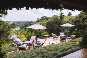 Een terras buiten met rieten meubilair en parasols, met uitzicht op een weelderig landschap met een rivier en heuvels.
