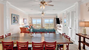A spacious dining area with a wooden table and red chairs, a large window with a view, and a ceiling fan. - Beachfront Bliss | Heated Private Pool | Pet Friendly (Gulf Shores)