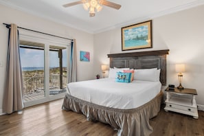 A bedroom with a large bed, a nightstand, a painting, and a view of the beach through a sliding door.