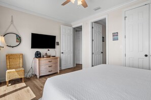 A bedroom with a large bed, a television mounted on the wall, a wicker chair, a dresser, and two doors leading to other rooms.