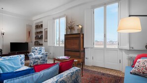 Un soggiorno con divano, poltrona e televisore. Si nota una libreria, un mobile in legno e una finestra con vista.
