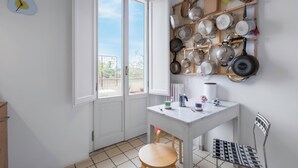 Una cucina con un tavolo bianco, uno sgabello di legno e una sedia in metallo. Sono presenti due finestre e una porta.