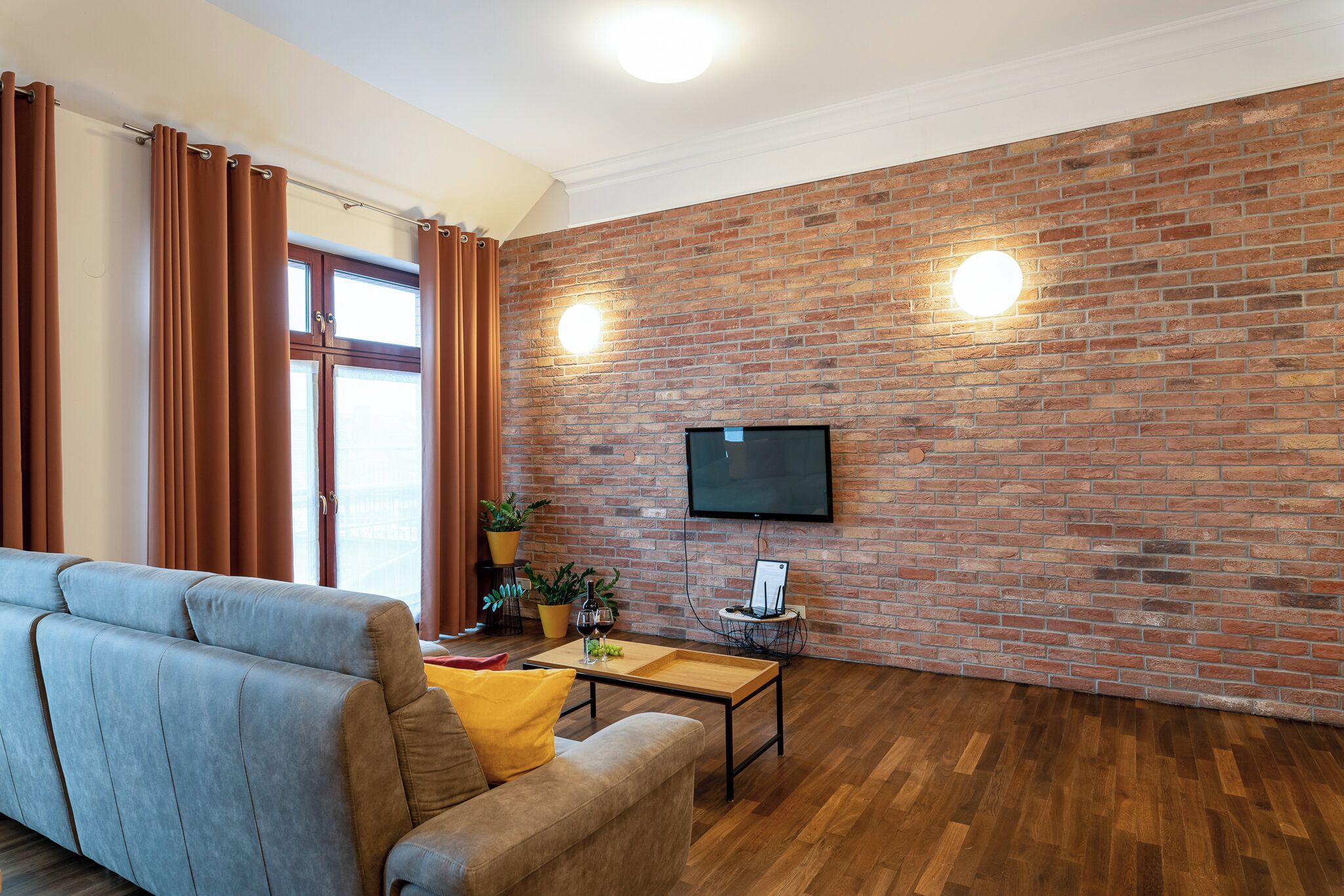 A living room with a brick wall, a flat-screen TV, a sofa, a coffee table, and a window with curtains.