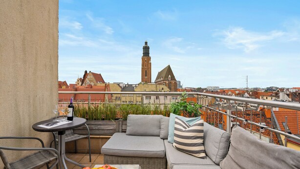 A balcony with wicker furniture, a table with a bottle and glasses, and a bowl of fruit. A cityscape with a prominent tower is visible in the background. - AS HOME Apartamenty Rynek 50 (Wroclaw)