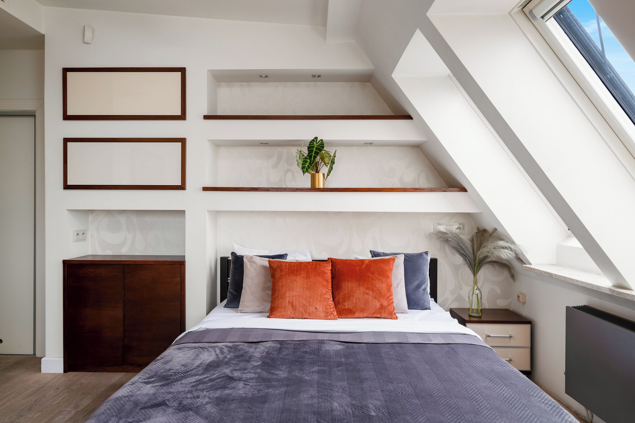 A modern bedroom with a sloped ceiling, built-in shelves, a bed with a grey and orange bedspread, and a small bedside table with a vase.