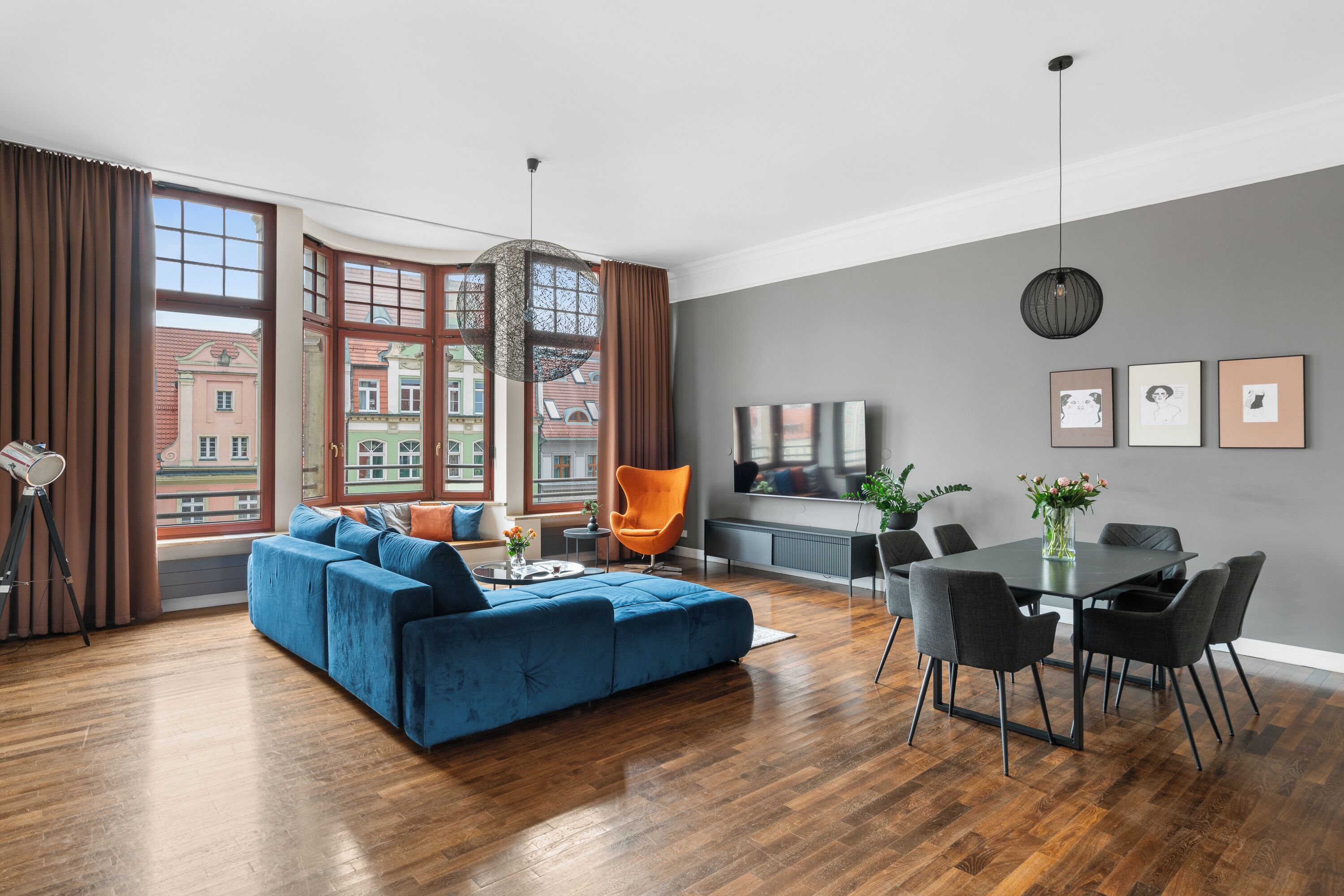 A modern living room with a blue sofa, dining table, and chairs. There are large windows with curtains, a television, and framed pictures on the wall.