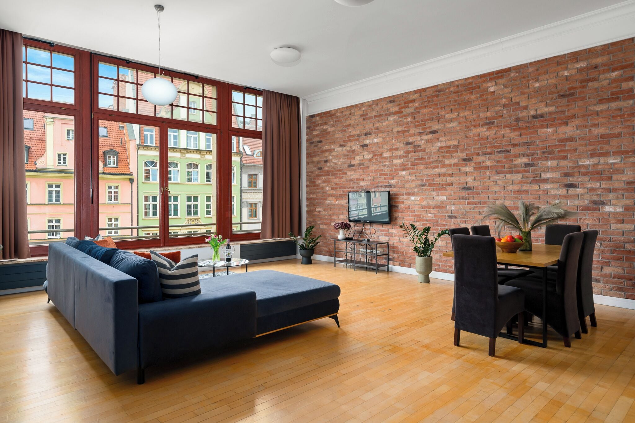 A spacious living room with a large window, a brick wall, a sofa, a dining table with chairs, and a flat-screen TV.