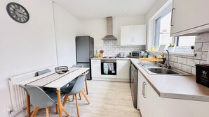 A compact kitchen with white cabinets, a built-in oven, a microwave, and a sink. There is a dining table with chairs and a clock on the wall. - Pengam House (Cardiff)