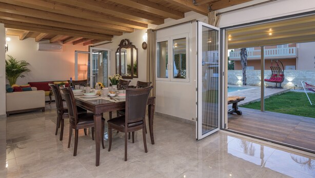 A dining area with a wooden table and chairs, a sofa, and a glass door leading to an outdoor patio with a pool and lounge chairs. - Garden Villa - bikes, private pool, garden family home (Crete)