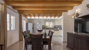 A spacious dining area with a wooden ceiling, a large dining table set for four, and a kitchen in the background. - Garden Villa - bikes, private pool, garden family home (Crete)