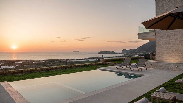 A modern outdoor pool area with lounge chairs and a view of the ocean at sunset. - GG Villas Falassarna (Chania)