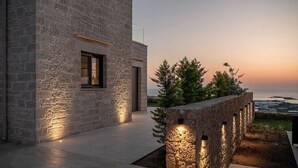 A stone building with a large window and a door, a stone wall with embedded lights, and a view of the sea at sunset. - GG Villas Falassarna (Chania)