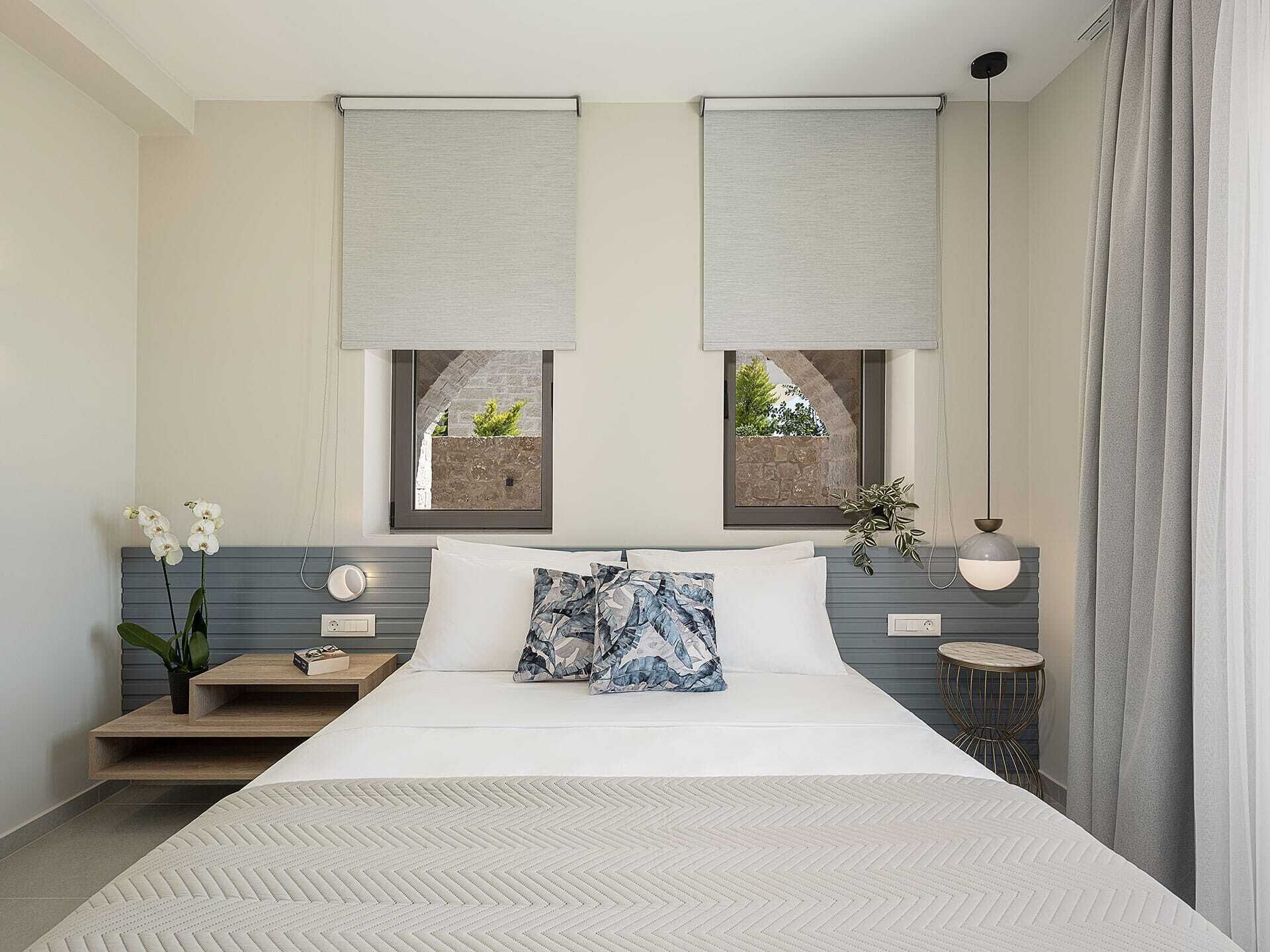 A modern bedroom with a large bed, two windows with view boxes, a bedside table with a plant, and a hanging light fixture.