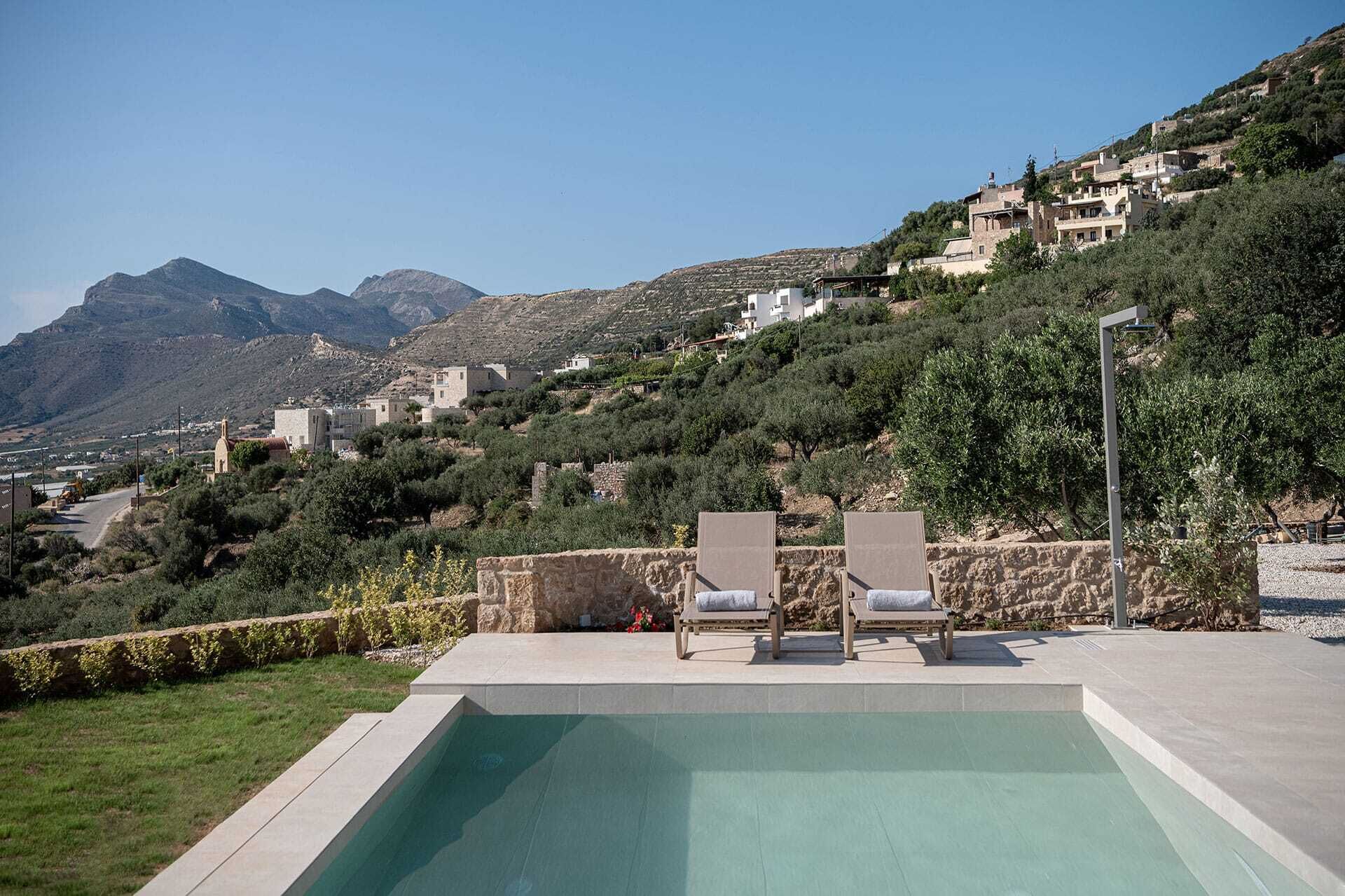 Ein Pool mit zwei Liegestühlen, umgeben von einer Steinmauer, mit Blick auf einen Hügel mit Gebäuden und Bergen.