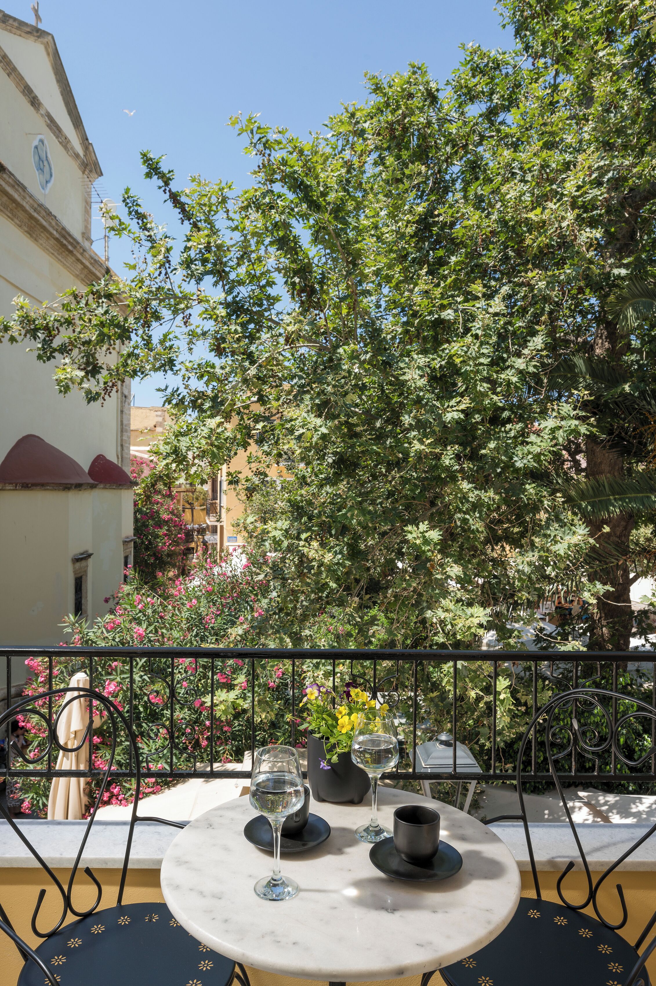 Un tavolo rotondo con due bicchieri da vino e una tazza, su uno sfondo con un albero e un edificio.