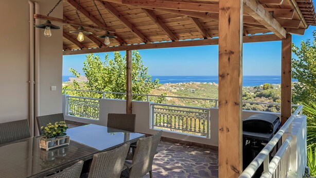 A covered outdoor patio with a table and chairs, a grill, and a view of the sea.