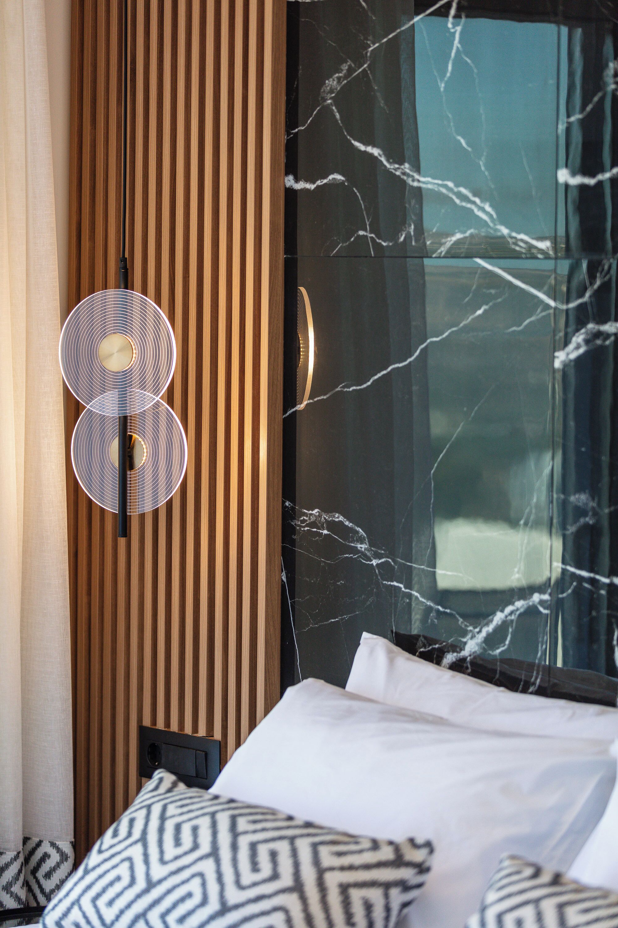 A hotel room with a bed, patterned pillows, a wooden panel wall, and a marble-framed window.
