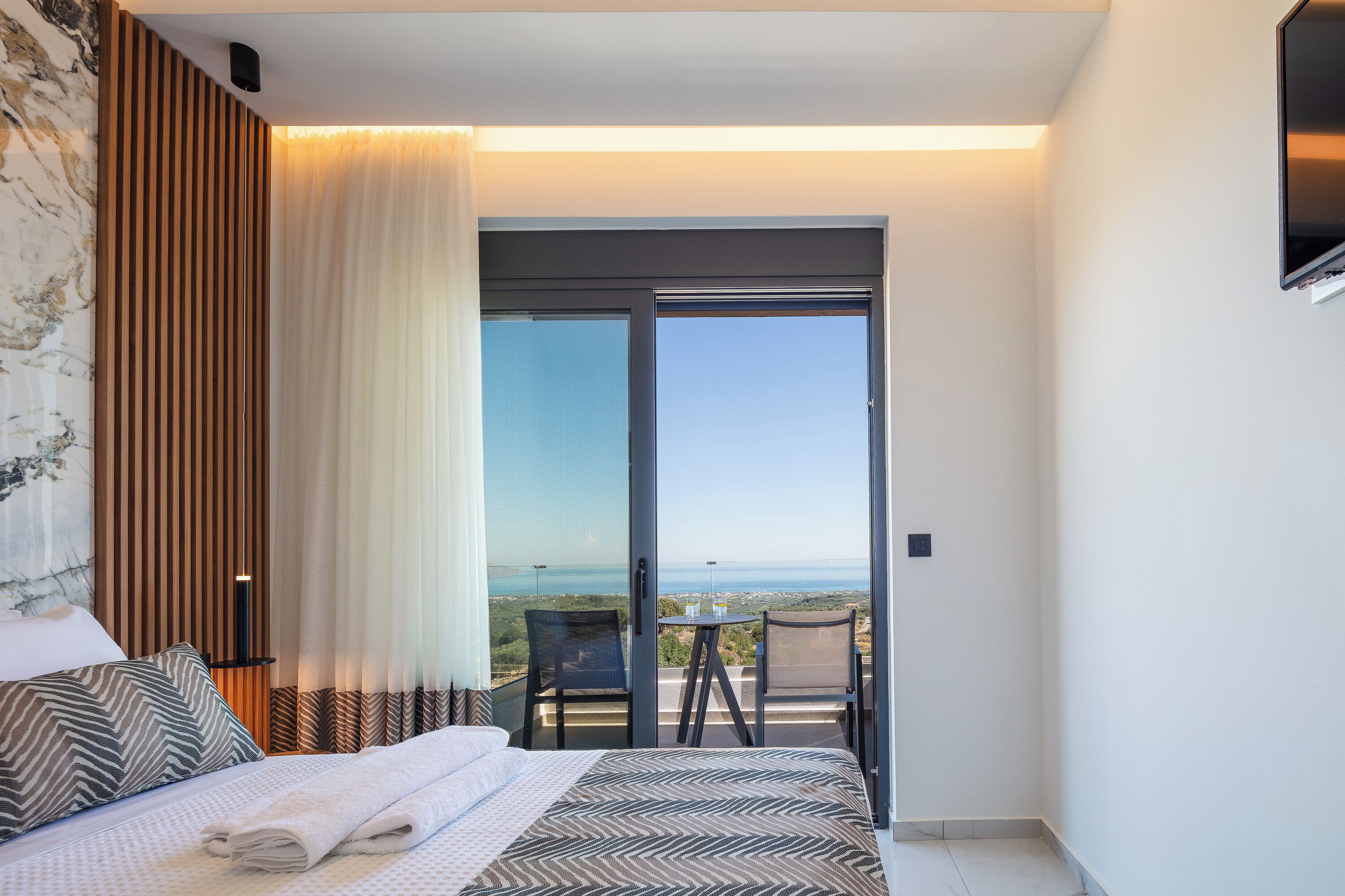 A bedroom with a bed, a view through a sliding glass door, a small table with chairs, and a TV mounted on the wall.