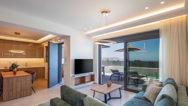 A modern living room with a sofa, coffee table, and TV. There is a kitchen area with cabinets and a dining table. The room has large windows and a view of the outdoors. - Horizon West Villa │Private Pool, Secluded Luxury Modern Villa (Chania)